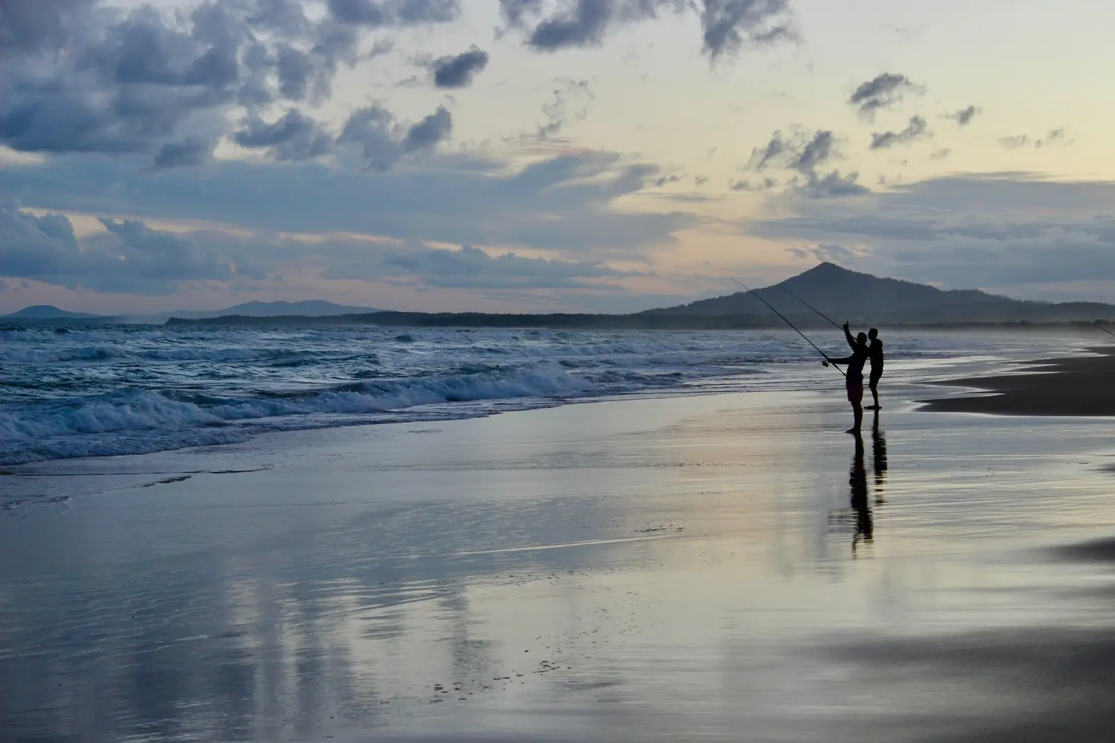 Two people fishing on a beach at sunset, with a mountain on the horizon